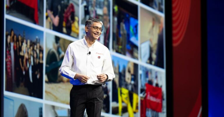 Manish Kumar, CEO da SolidWorks, está em um palco apresentando-se diante de um grande telão que exibe um mosaico de imagens diversas. Ele veste uma camisa branca com o logotipo da empresa e um crachá comemorativo de 30 anos. Manish Kumar sorri enquanto fala, transmitindo um tom confiante e envolvente. O fundo inclui tons de azul e vermelho, sugerindo um ambiente corporativo e tecnológico
