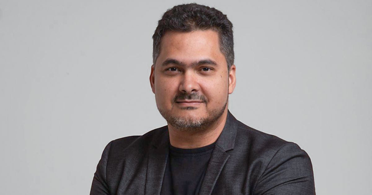 Foto de um homem de pele clara, barba curta e cabelo escuro levemente ondulado. Ele veste um blazer preto sobre uma camisa preta e tem uma expressão confiante e amigável. Seus braços estão cruzados e o fundo é um cinza neutro, conferindo um aspecto profissional à imagem (token)