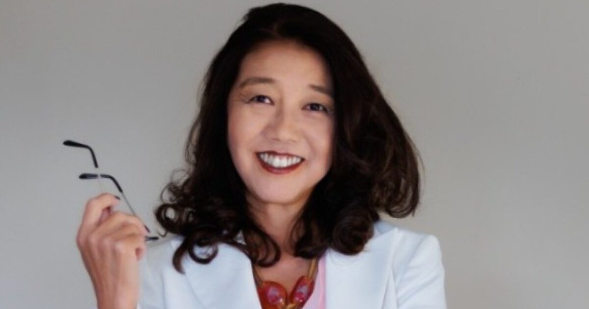 Nancy Abe sorridente de cabelos castanhos longos e ondulados, vestindo um blazer branco sobre uma blusa rosa e um colar de pedras vermelhas. Ela segura um par de óculos na mão direita e olha diretamente para a câmera com expressão confiante. O fundo é neutro e cinza claro.