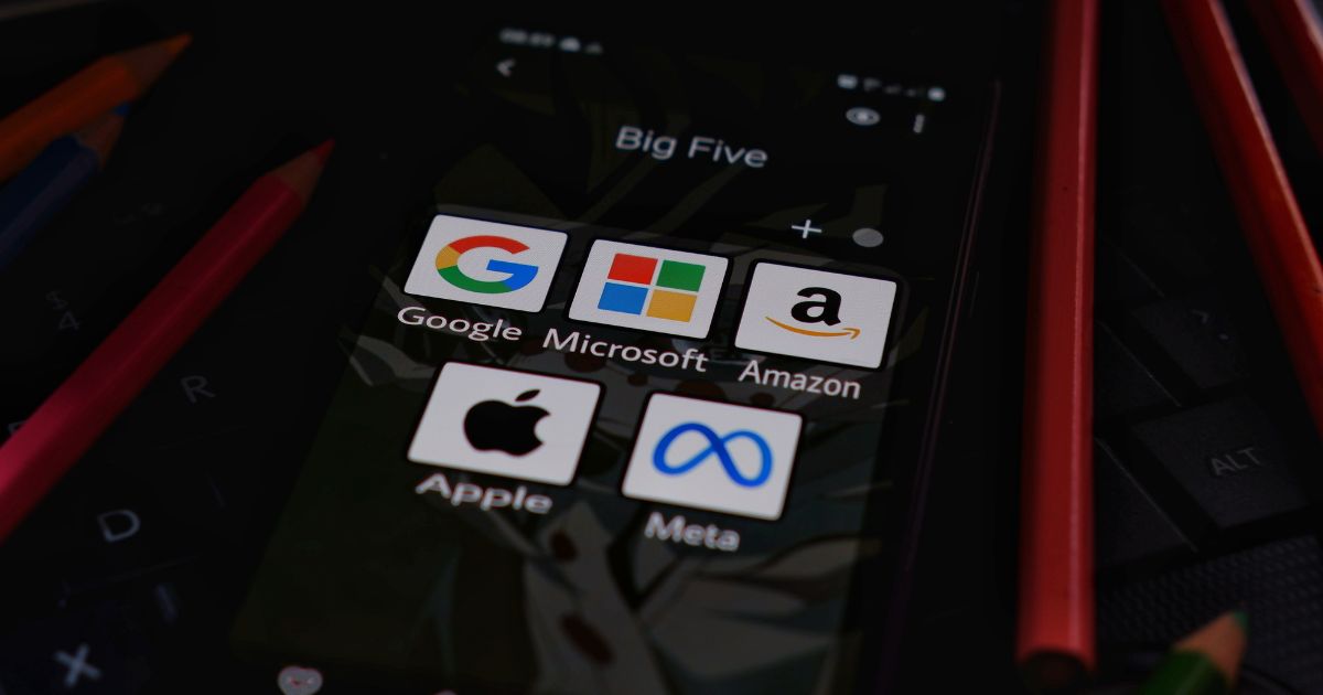 Ecrã de um smartphone exibindo uma pasta chamada 'Big Five', contendo os ícones das cinco maiores empresas de tecnologia: Google (com o logótipo colorido da letra G), Microsoft (logótipo quadrado multicolorido), Amazon (logótipo preto com uma seta amarela), Apple (logótipo da maçã mordida em preto) e Meta (logótipo azul semelhante a um infinito, ações). O telemóvel está sobre um teclado de computador e há lápis coloridos ao redor da cena.
