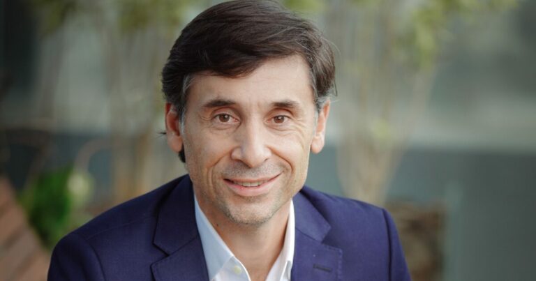 Retrato de Willian Pimentel, vice-presidente da Freshworks na América Latina. Ele está sentado ao ar livre, vestindo um terno azul e camisa branca, sorrindo para a câmera. A imagem transmite uma atmosfera profissional e acessível. Software, IA