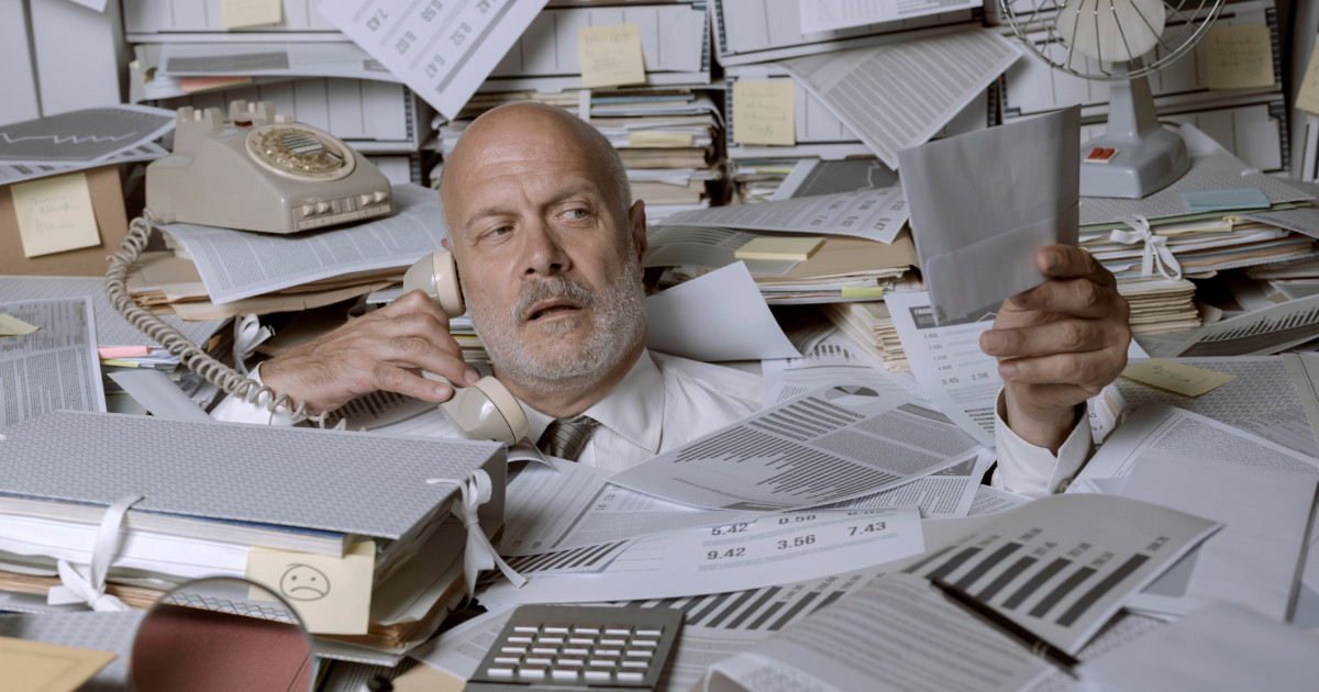 A imagem mostra um homem de meia-idade, de aparência exausta, vestindo uma camisa branca e gravata, cercado por uma enorme quantidade de papéis, documentos, gráficos e arquivos. Ele está ao telefone com uma expressão de cansaço e confusão, enquanto segura um envelope ou ficha com a outra mão. O ambiente parece ser um escritório caótico, com uma mesa e prateleiras desorganizadas, lotadas de papéis, pastas e notas adesivas. Ao fundo, um telefone antigo, um ventilador e mais pilhas de documentos reforçam a sensação de desordem extrema (caos)
