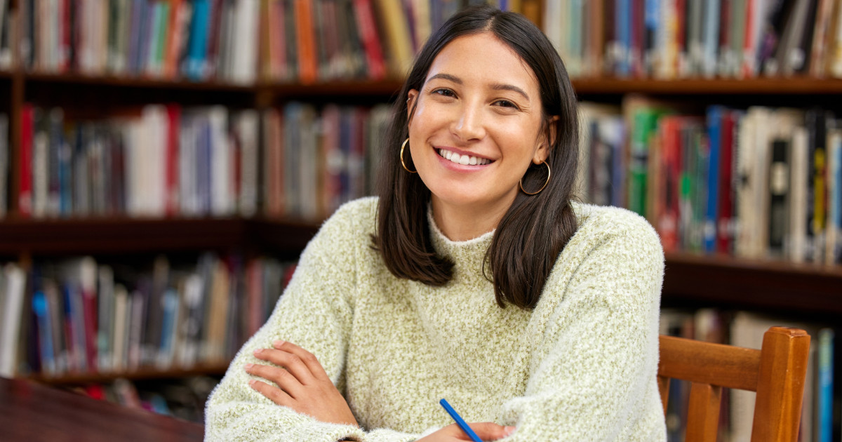 A imagem mostra uma jovem estudante sorrindo enquanto está sentada em uma biblioteca. Ela usa um suéter claro e segura uma caneta azul, aparentando estar em um momento de estudo ou escrita. Ao fundo, há estantes repletas de livros, criando um ambiente acadêmico e acolhedor (exige)