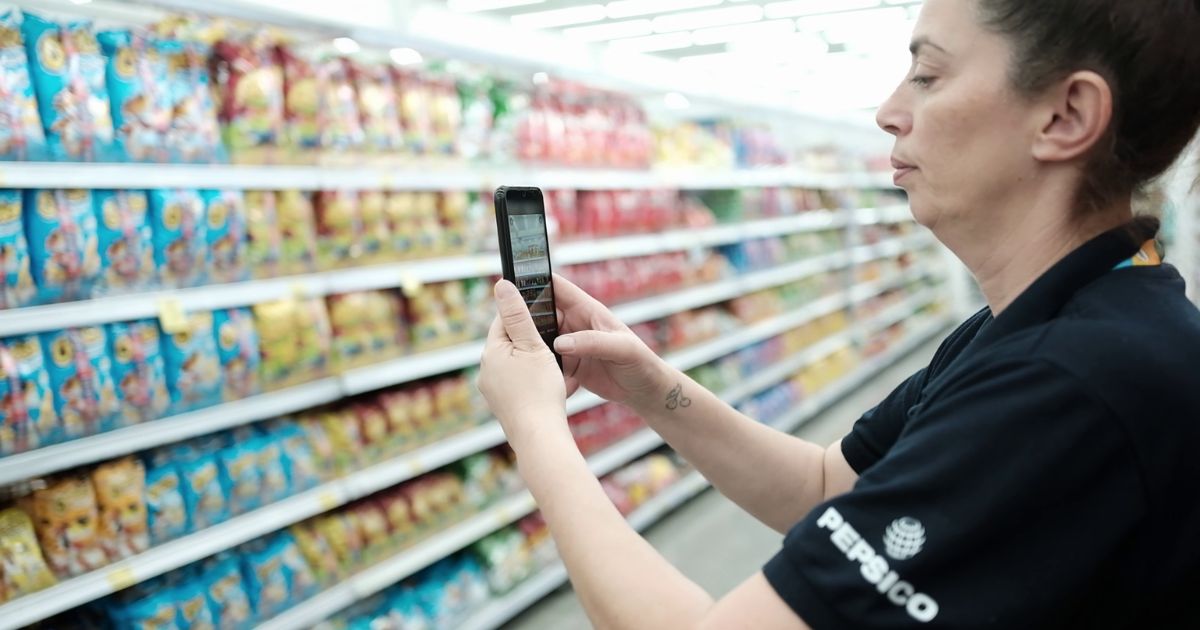 A imagem mostra uma promotora de vendas em um ponto de venda (PDV) de supermercado, vestindo uma camisa preta com o logo da PepsiCo. Ela segura um smartphone e parece estar registrando ou verificando informações sobre os produtos dispostos nas prateleiras, que contêm pacotes coloridos de salgadinhos e snacks. O ambiente é bem iluminado, e a organização dos produtos sugere um processo de controle de estoque, precificação ou merchandising (PDVs)