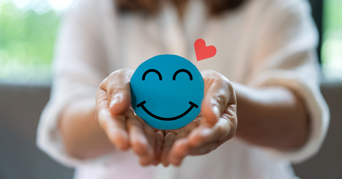 A imagem mostra uma pessoa segurando delicadamente um círculo azul com um rosto sorridente desenhado, representando positividade e bem-estar. Acima, há um pequeno coração vermelho, reforçando a ideia de cuidado e saúde mental. O fundo é desfocado, dando destaque às mãos e ao símbolo de felicidade