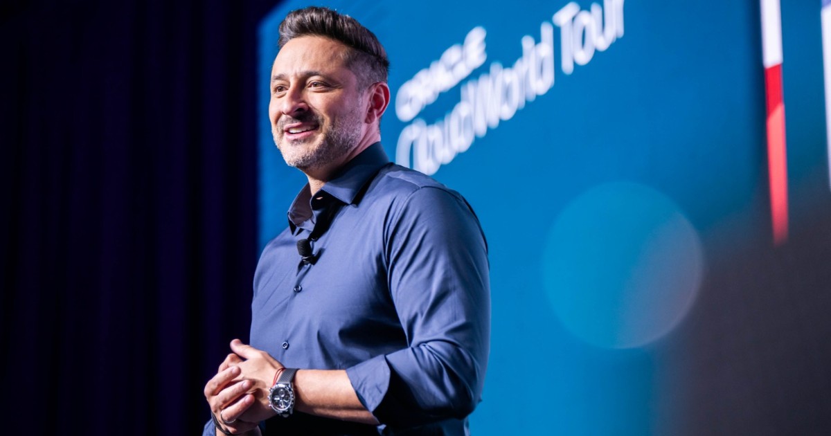 Alexandre Maioral, presidente da Oracle do Brasil, está em pé no palco durante um evento. Ele sorri enquanto segura um controle nas mãos. Ao fundo, há uma grande tela azul com seu nome e cargo exibidos: "Alexandre Maioral, Presidente, Oracle do Brasil".