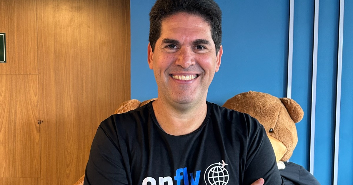 Homem sorridente de cabelos escuros, vestindo uma camiseta preta com o logotipo da empresa "Onfly", está de braços cruzados em frente a uma parede azul e uma parede de madeira. Atrás dele, há um grande urso de pelúcia marrom. A imagem mostra Christian Machado, novo CTO da Onfly