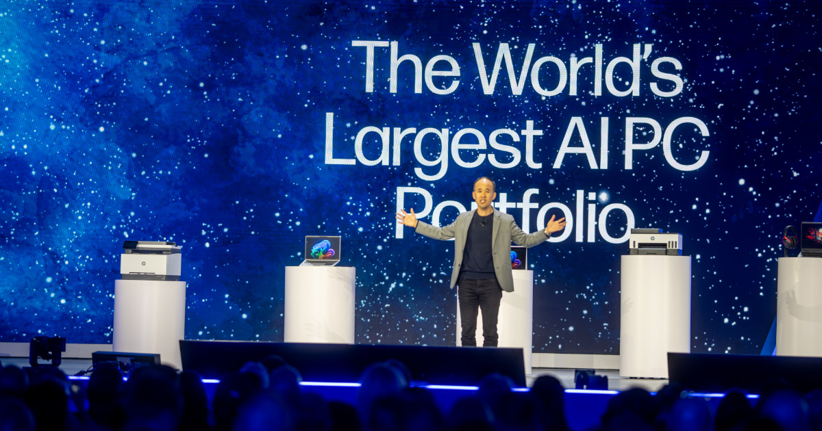 A imagem mostra Alex Cho, presidente de Personal Systems da HP, no palco durante um evento da HP Amplify. Ele está apresentando novos produtos da linha de PCs com inteligência artificial, com um grande telão ao fundo exibindo a frase "The World's Largest AI PC Portfolio" ("O maior portfólio de PCs com IA do mundo"). No palco, há pedestais com notebooks, impressoras e outros dispositivos da HP. A plateia aparece na parte inferior da imagem, assistindo à apresentação