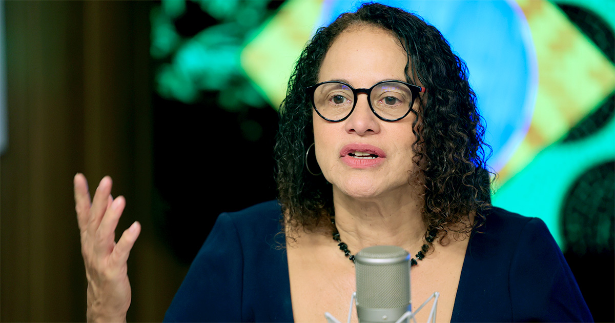 A imagem mostra Luciana Santos, ministra de Ciência, Tecnologia e Inovação do Brasil, falando em um microfone. Ela veste uma blusa azul escura, usa óculos de armação preta e tem cabelos cacheados e soltos. Sua expressão é séria e envolvente, com uma das mãos levantadas enquanto fala. O fundo desfocado exibe as cores da bandeira do Brasil, sugerindo um ambiente institucional ou governamental