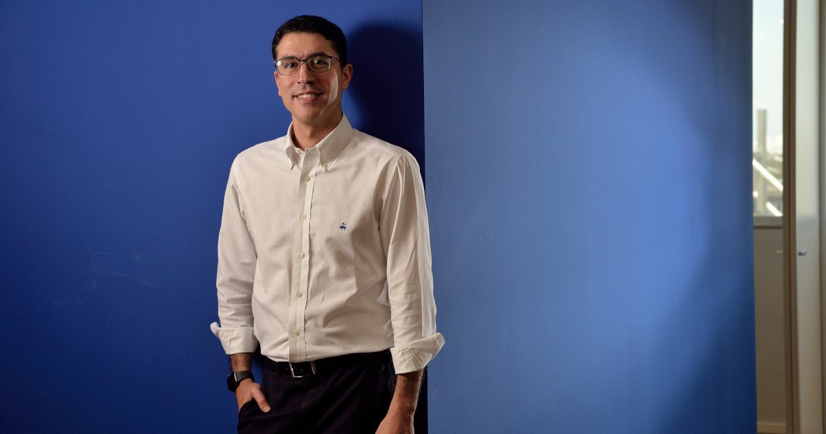 Imagem de Marcelo Mearim, CEO da Sofya, em pé diante de uma parede azul. Ele está sorrindo, veste uma camisa social branca de manga comprida com o logotipo discreto da marca na altura do peito e calça escura. Usa óculos e um relógio no pulso esquerdo. Ao fundo, há uma janela com vista externa parcialmente visível (saúde)