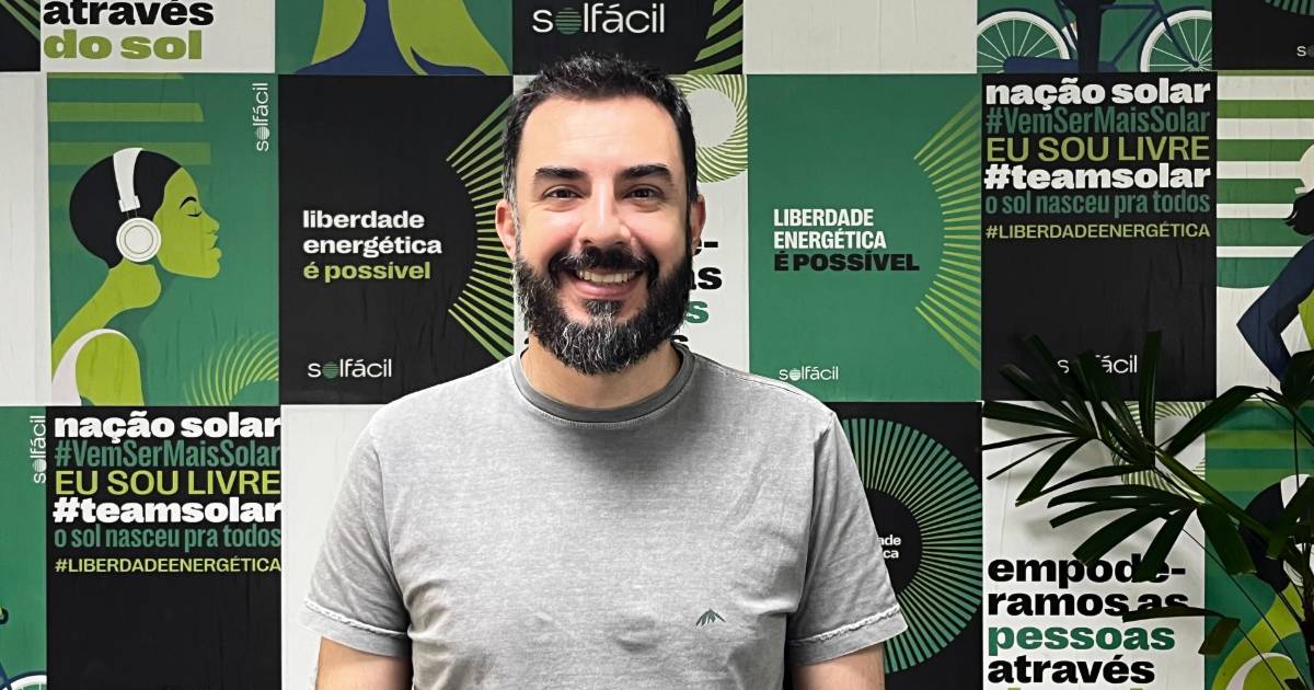 Homem de barba e cabelo escuros, usando camiseta cinza clara, sorri em frente a uma parede com ilustrações e frases relacionadas à energia solar. O fundo traz a identidade visual da empresa Solfácil, com mensagens como “liberdade energética é possível” e “#teamsolar”. Na imagem, está Mateus Neves, que foi anunciado como o novo CTO da Solfácil