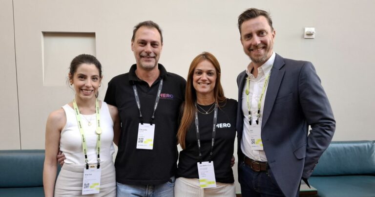 Quatro pessoas posam juntas para uma foto em um ambiente interno com um fundo neutro e um sofá azul. Eles estão sorrindo e vestindo crachás de identificação em cordões pretos e amarelos. Duas pessoas estão vestindo camisetas pretas com a marca "HERO", enquanto as outras duas usam roupas mais formais — uma mulher de blusa branca e um homem de blazer azul. A atmosfera sugere um evento profissional ou conferência de negócios.