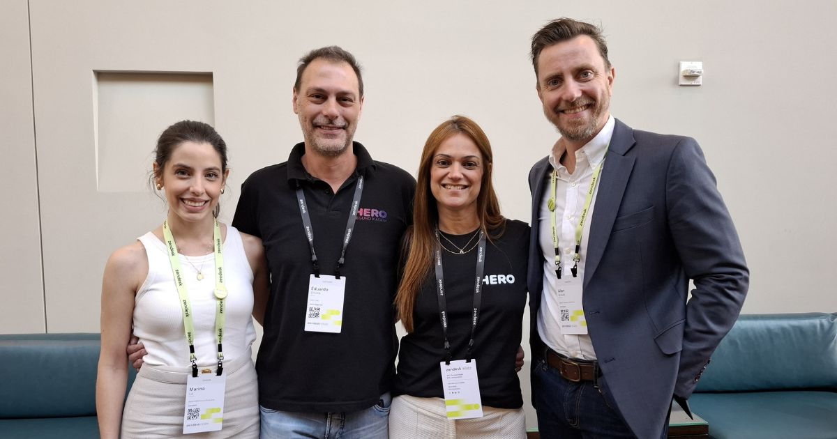 Quatro pessoas posam juntas para uma foto em um ambiente interno com um fundo neutro e um sofá azul. Eles estão sorrindo e vestindo crachás de identificação em cordões pretos e amarelos. Duas pessoas estão vestindo camisetas pretas com a marca "HERO", enquanto as outras duas usam roupas mais formais — uma mulher de blusa branca e um homem de blazer azul. A atmosfera sugere um evento profissional ou conferência de negócios.