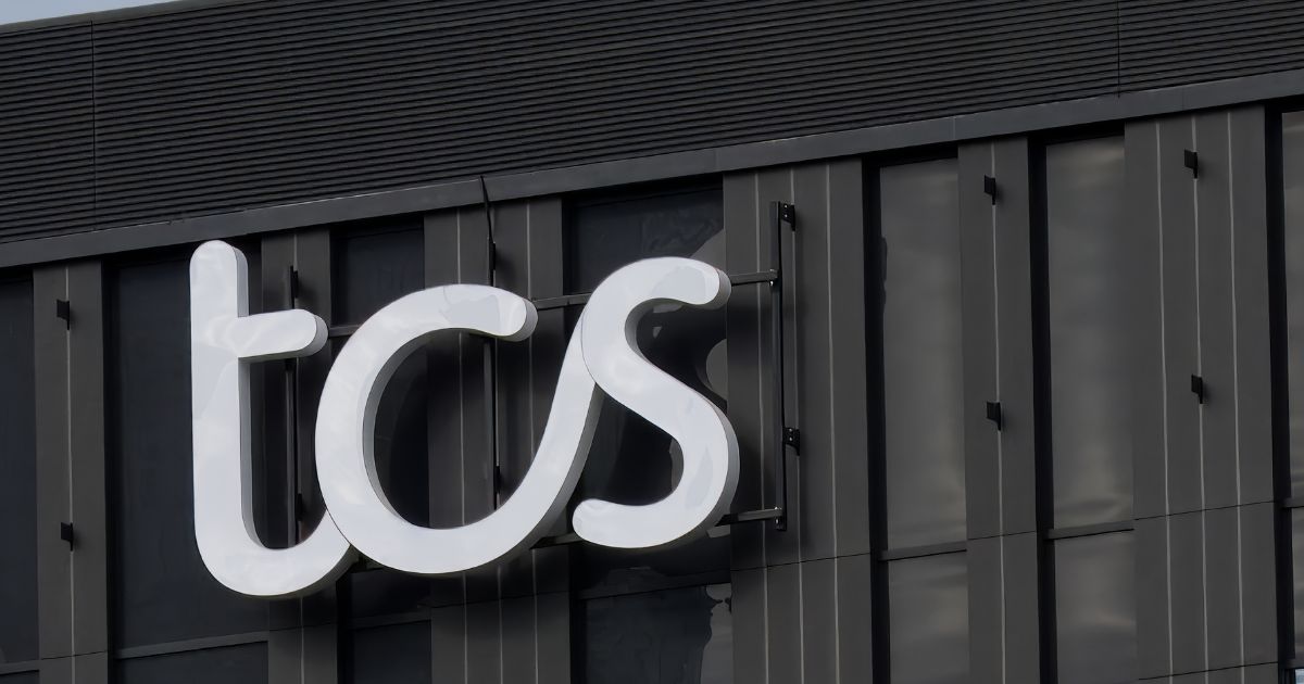 Um letreiro iluminado com as letras "tcs" em branco está fixado na fachada de um prédio moderno de cor escura. A estrutura do edifício apresenta janelas verticais e detalhes arquitetônicos minimalistas. O céu ao fundo está parcialmente nublado, refletindo sutilmente na superfície do letreiro (mulher)