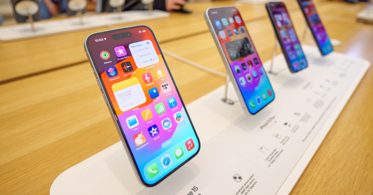 Uma fileira de iPhones 15 em exibição em uma loja da Apple, posicionados em suportes inclinados sobre uma mesa de madeira. O primeiro iPhone na frente está em destaque, exibindo uma tela colorida com diversos aplicativos e widgets. Ao fundo, outros modelos estão alinhados, levemente desfocados, enquanto alguns clientes podem ser vistos explorando o ambiente da loja