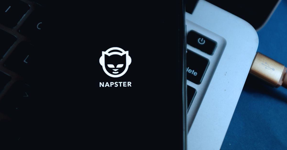 Uma fotografia mostra um smartphone com a tela ligada exibindo o logotipo do Napster, que consiste em um ícone de um gato estilizado usando fones de ouvido, acompanhado do nome "NAPSTER" em letras maiúsculas. O telefone está apoiado sobre um teclado de computador e uma parte de um laptop prateado é visível ao fundo, com um cabo USB dourado conectado a ele. O ambiente tem iluminação suave, criando um tom tecnológico e nostálgico.