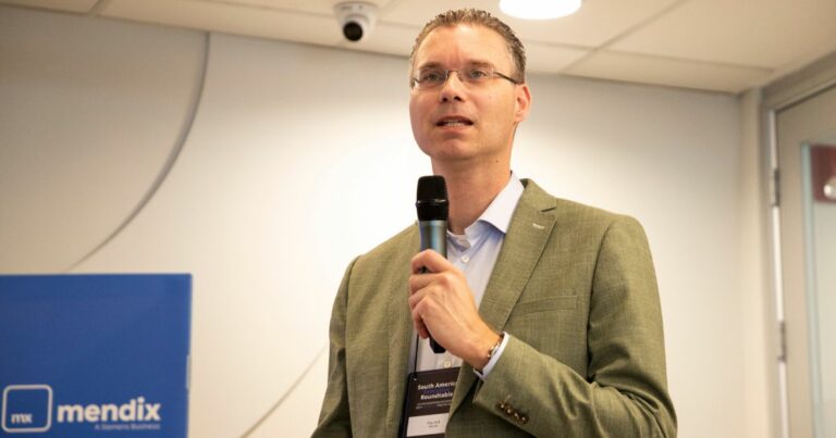 Um homem de óculos, vestido com um blazer verde-oliva e camisa social clara, está segurando um microfone enquanto fala para um público. Ele tem uma expressão confiante e profissional. Ao fundo, há uma parede branca com um logótipo azul da 'Mendix' e uma câmara de segurança no teto. O ambiente parece ser uma sala de conferências ou evento corporativo.