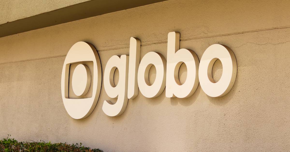 Logotipo da TV Globo fixado em uma parede externa de cor bege. O logo é tridimensional, em tom claro, com a palavra "globo" em letras minúsculas ao lado do símbolo característico da emissora — um círculo central dentro de uma forma ovalada, representando uma tela. A base da parede possui uma pequena vegetação (Zoho)