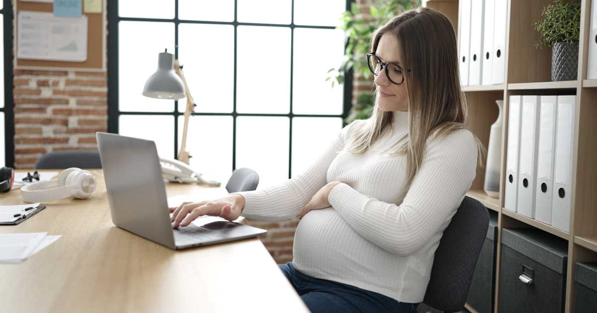 A imagem mostra uma mulher grávida trabalhando em um ambiente de escritório moderno e bem iluminado. Ela veste uma blusa branca de mangas longas e óculos, e está sentada em uma cadeira confortável, utilizando um laptop sobre a mesa. Com uma das mãos no teclado e a outra apoiada na barriga, ela demonstra concentração e tranquilidade. O espaço ao redor tem prateleiras com pastas organizadas, plantas e uma grande janela com estrutura de metal, permitindo a entrada de luz natural.