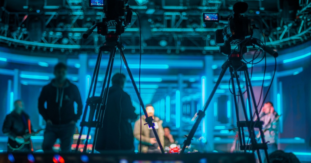 Estúdio de gravação com ambiente futurista iluminado por luzes azuis neon. No primeiro plano, duas câmeras profissionais em tripés capturam a cena. Ao fundo, pessoas aparecem desfocadas, incluindo um músico tocando guitarra e uma equipe de produção. O cenário apresenta um design moderno com painéis iluminados, criando uma sensação de profundidade e tecnologia, Huawei
