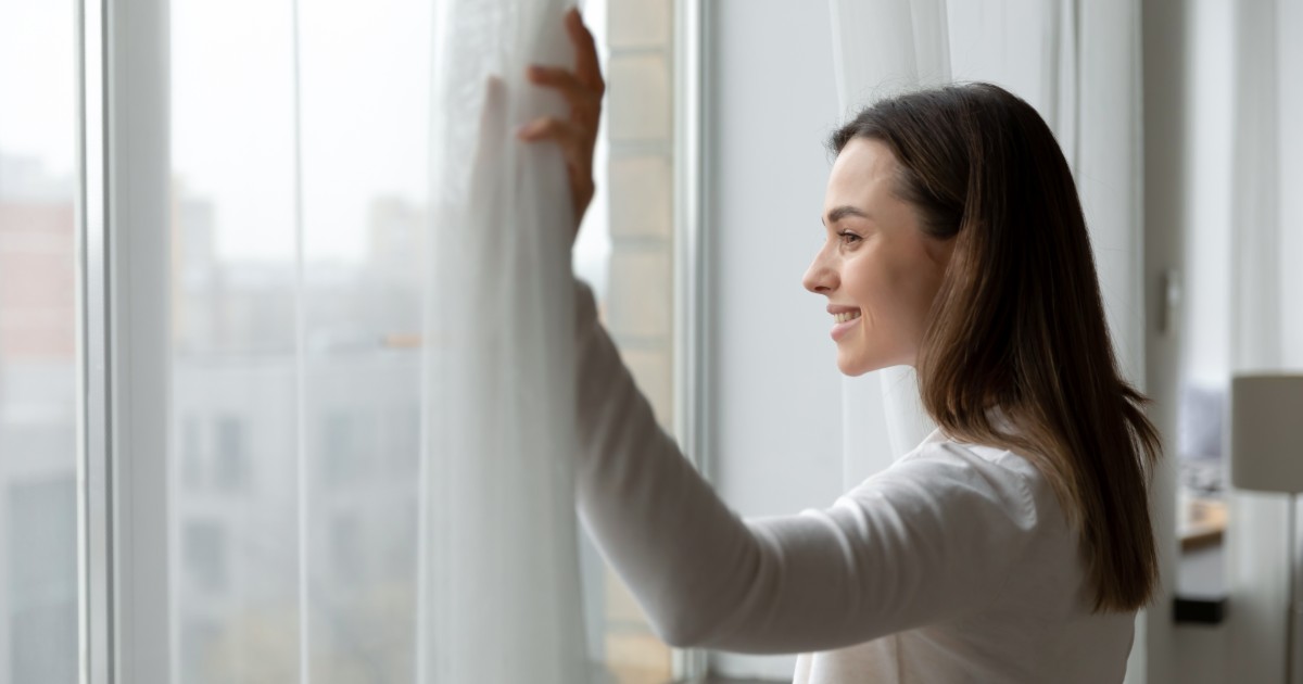 A imagem mostra uma mulher sorridente abrindo cortinas brancas em uma janela grande, permitindo a entrada de luz natural. Ela veste uma blusa clara e parece estar em um ambiente confortável e moderno. O fundo mostra um cenário urbano levemente desfocado. A cena transmite uma sensação de frescor, bem-estar e início de um novo dia (começando o dia)