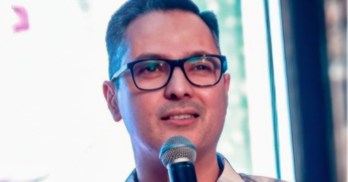 Eronides Junior, novo Chief Revenue Officer (CRO) da SoftwareOne, aparece segurando um microfone e sorrindo, usando óculos de grau e camisa clara, com fundo desfocado e iluminação suave.