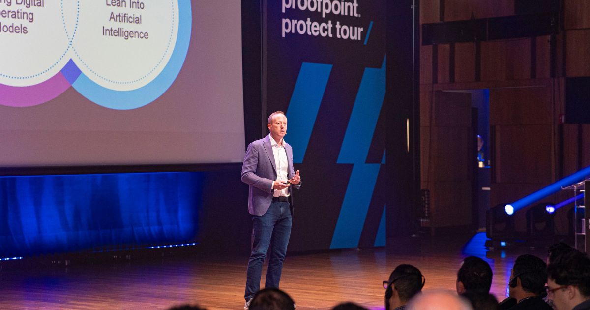 Homem a fazer uma apresentação num palco durante o evento "Proofpoint Protect Tour". Ele está vestido com blazer, camisa branca e jeans, segurando um comando. À sua esquerda há um ecrã com gráficos e textos relacionados com inteligência artificial. O público está sentado a assistir, e o ambiente é profissional e moderno, com iluminação azul. Nate Chessin, Vice-presidente sênior de engenharia de sistemas mundiais da Proofpoint