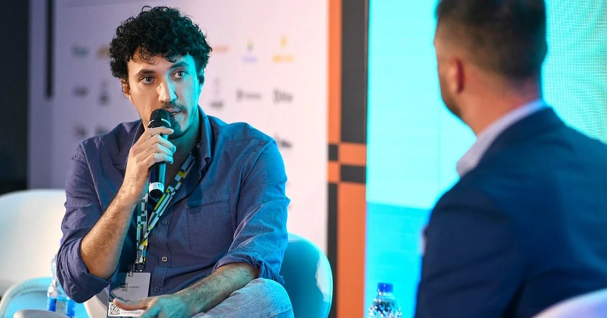 Dois homens participam numa conversa durante um painel de conferência. O homem em primeiro plano (à esquerda) está a falar ao microfone, vestindo uma camisa azul e crachá ao pescoço. Está sentado e com expressão atenta. À direita, parcialmente desfocado, vê-se outro homem de costas a ouvi-lo. O fundo mostra um ecrã azul claro e um painel com logótipos de patrocinadores ou marcas (Bitshopp)