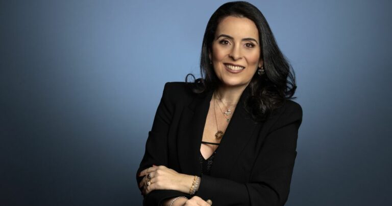 Uma mulher de cabelos longos e escuros posa sorrindo para a câmera contra um fundo azul. Ela usa um blazer preto elegante sobre uma blusa com decote e vários acessórios, incluindo colares, anéis e pulseiras. Seu olhar é confiante e amigável, e sua postura transmite profissionalismo e simpatia. Claudia Muchaluat