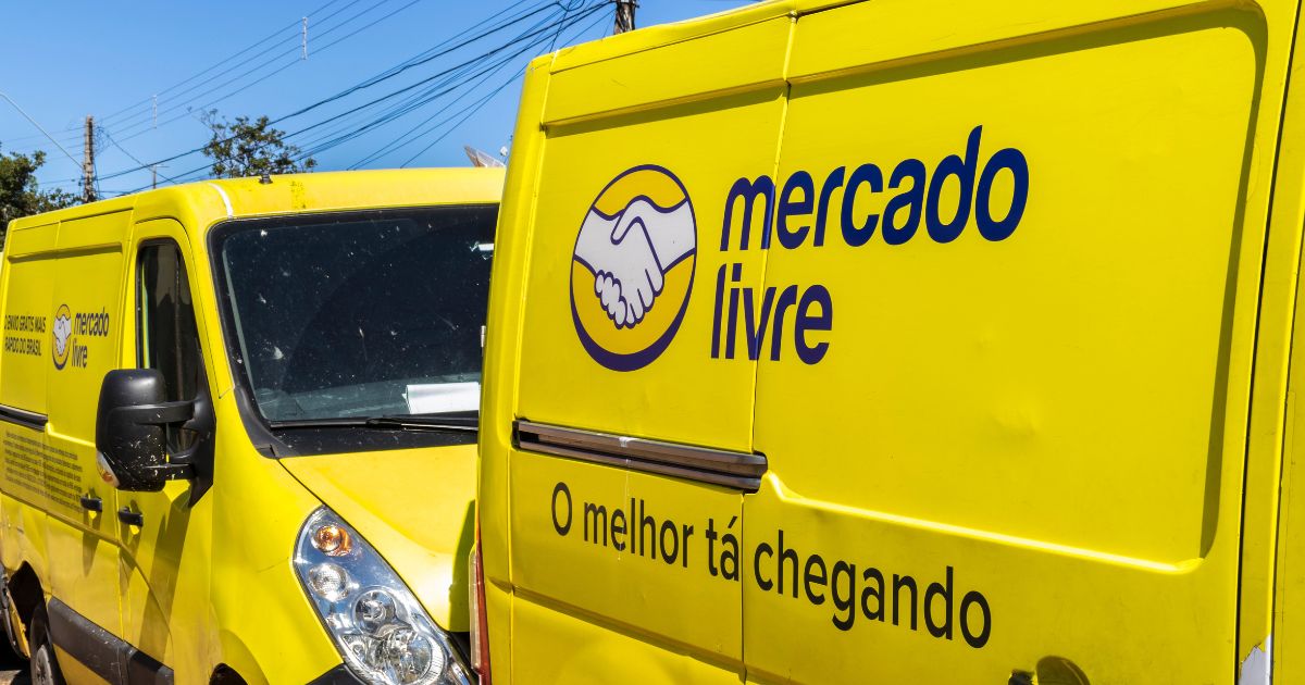 Fotografia de duas carrinhas amarelas da empresa Mercado Livre estacionadas lado a lado. As viaturas exibem o logótipo da marca — um aperto de mãos dentro de um círculo — e o nome 'mercado livre' em letras azuis. Numa das portas está escrita a frase promocional 'O melhor tá chegando'. Ao fundo, veem-se postes e fios elétricos, sugerindo um ambiente urbano.