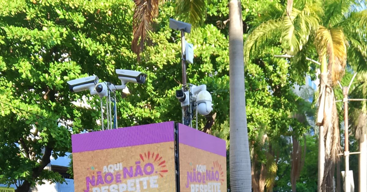 Imagem de um totem de segurança pública instalado em área externa com várias câmeras de vigilância voltadas em diferentes direções. O totem exibe o aviso colorido: 'AQUI NÃO É NÃO. RESPEITE', indicando uma campanha de conscientização contra o assédio, possivelmente em evento público. Ao fundo, há árvores tropicais e postes, sugerindo um ambiente urbano arborizado.