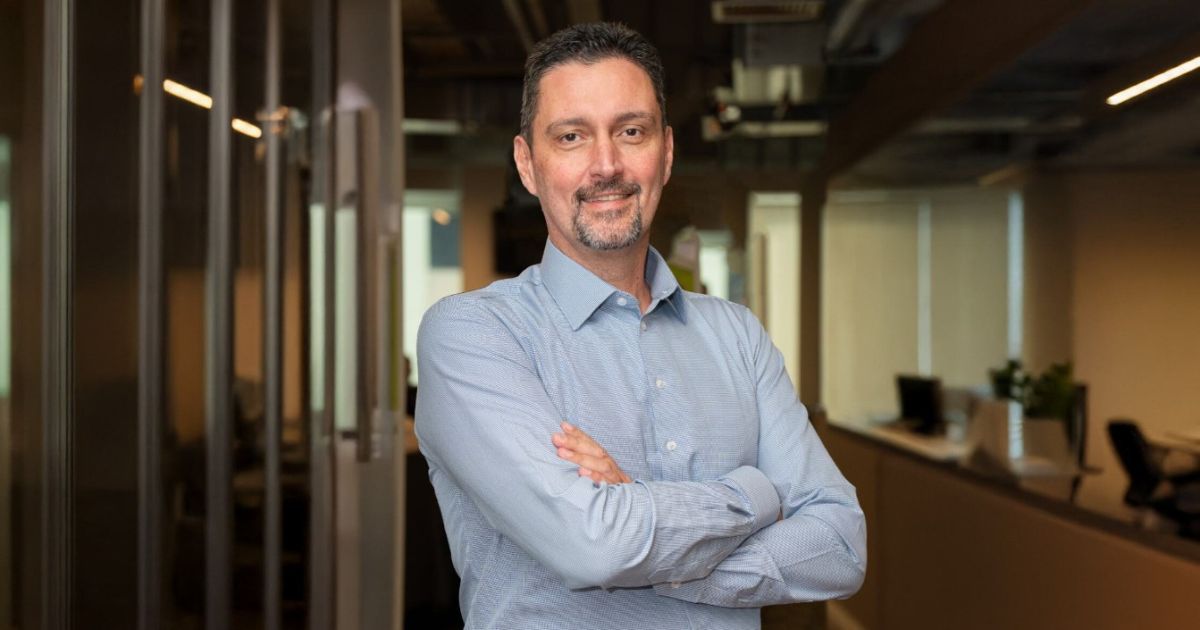 Homem de meia-idade com cabelo curto escuro e barba bem aparada, veste uma camisa azul clara e está de braços cruzados, sorrindo com expressão confiante. Ele está num ambiente de escritório moderno com paredes de vidro e luz natural a entrar pelas janelas ao fundo. O espaço é iluminado e profissional, com secretárias e cadeiras visíveis ao fundo.