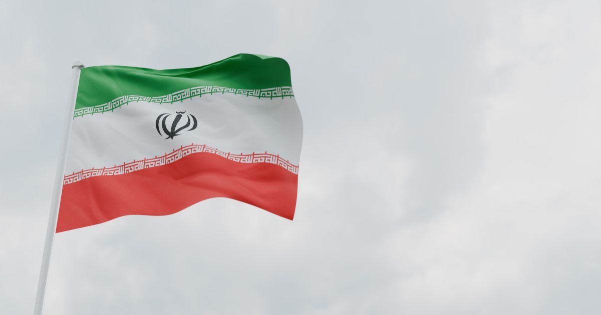 Imagem de uma bandeira do Irã tremulando ao vento contra um céu nublado. A bandeira tem três faixas horizontais nas cores verde, branca e vermelha, com inscrições em árabe estilizadas nas divisões entre as cores. No centro da faixa branca está o emblema nacional do Irã, em vermelho escuro.