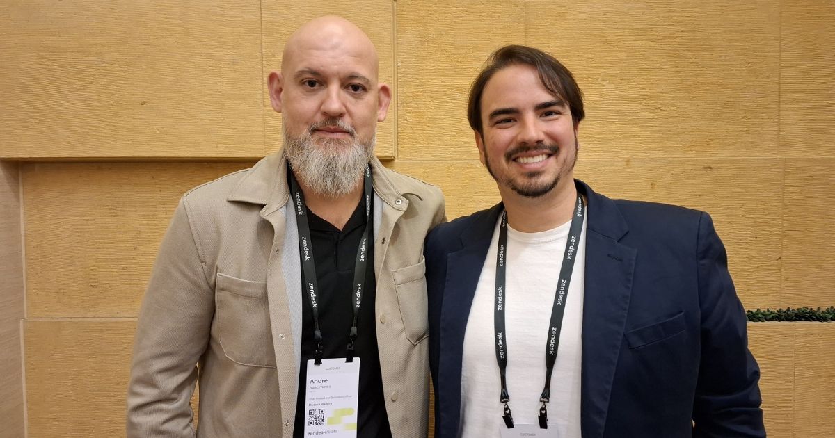 Dois homens posam juntos em um evento. O homem à esquerda tem barba longa e grisalha, cabeça raspada e veste uma jaqueta bege. O homem à direita tem cabelo escuro, curto e um sorriso no rosto, vestindo um blazer azul-marinho e uma camiseta branca. Ambos usam crachás pretos com a marca "Zendesk" e estão em um ambiente de conferência, com uma parede texturizada bege ao fundo. (MadeiraMadeira)