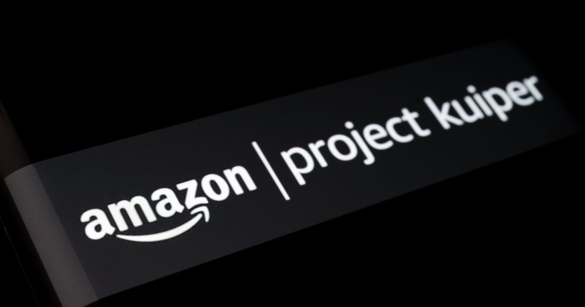 Imagem em close-up da tela de um dispositivo eletrônico mostrando o logotipo da Amazon ao lado do nome 'Project Kuiper' em letras brancas sobre um fundo preto. A imagem faz referência ao projeto da Amazon voltado para o lançamento de satélites e expansão de internet de banda larga ao redor do mundo.