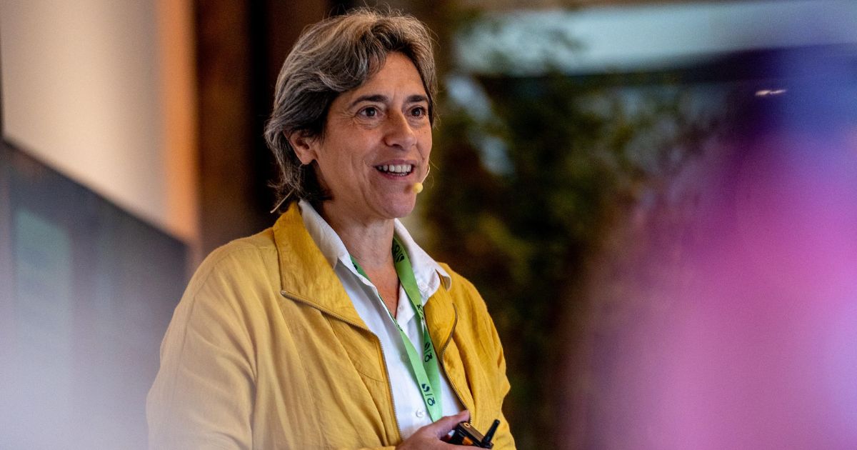 Mulher de cabelos grisalhos curtos, usando casaco amarelo sobre camisa branca, sorrindo enquanto fala com um microfone de lapela preso à face. Ela segura um controle remoto e usa um crachá verde no pescoço. Ao fundo, o ambiente está desfocado, sugerindo um espaço interno com iluminação suave. A mulher na imagem é Carla Tieppo, neurocientista