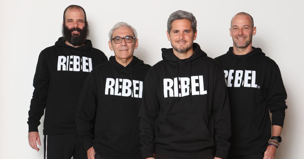 Imagem de quatro homens adultos posando lado a lado, sorridentes, diante de um fundo branco. Todos estão usando moletons pretos com a palavra 'REBEL' escrita em letras grandes e brancas na frente, sugerindo que fazem parte da mesma equipe, empresa ou campanha.