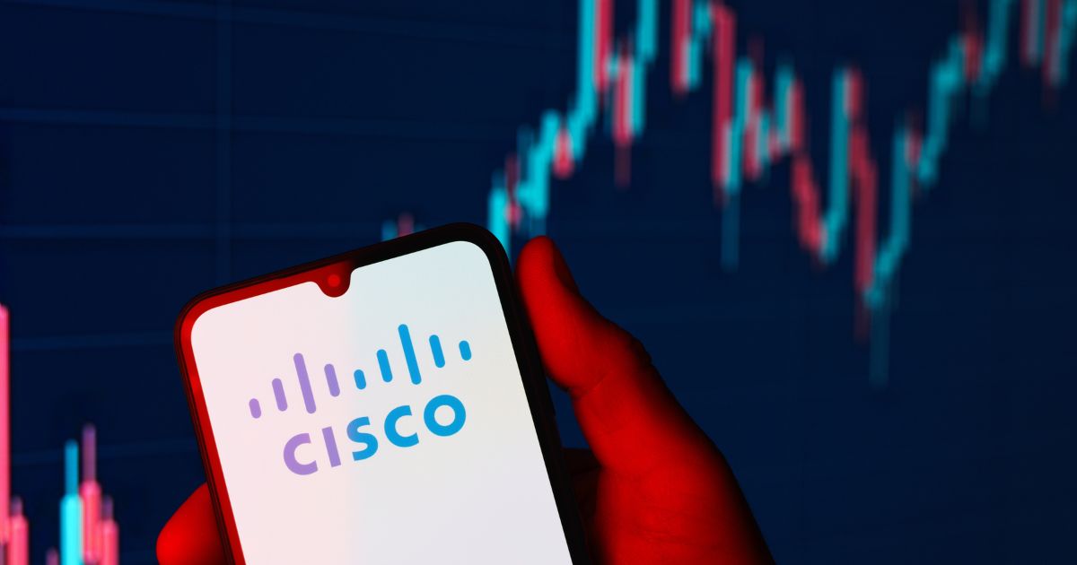 Uma mão segura um smartphone que exibe o logotipo da Cisco na tela. Ao fundo, há um gráfico de mercado financeiro com barras e linhas em tons de azul e vermelho, sugerindo análise de dados ou desempenho financeiro da empresa. A iluminação da cena cria um efeito dramático, com um tom avermelhado destacando a mão e o dispositivo.