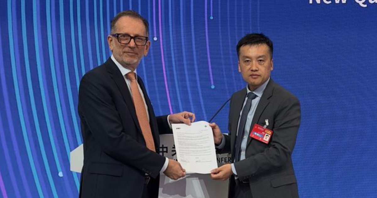 Rodolfo Fücher, vice-presidente do conselho da Abes, e Feng Bo, representante da ZGC International, posam ao centro da imagem enquanto seguram juntos uma carta de intenções assinada para a criação do ‘China Desk’ no Brasil. Ambos usam ternos formais; Fücher veste gravata laranja e óculos, enquanto Feng Bo tem crachá vermelho preso ao paletó. Ao fundo, há um painel azul com linhas curvas em tons de azul e roxo, com parte do texto “New Q…” visível, indicando o ambiente de uma conferência ou evento formal.