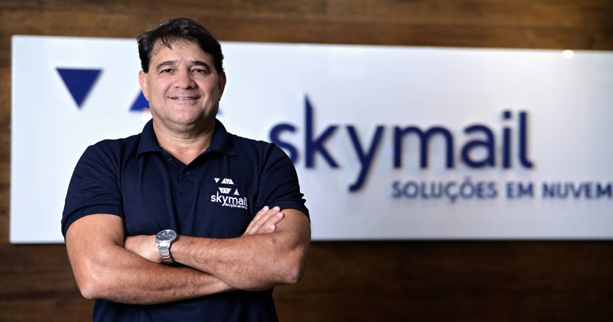 Paulo Lima, CEO da Skymail, posando de braços cruzados e sorrindo. Ele veste uma camisa polo azul-marinho com o logotipo da Skymail e está em frente a um painel com a marca da empresa e o slogan 'Soluções em Nuvem'