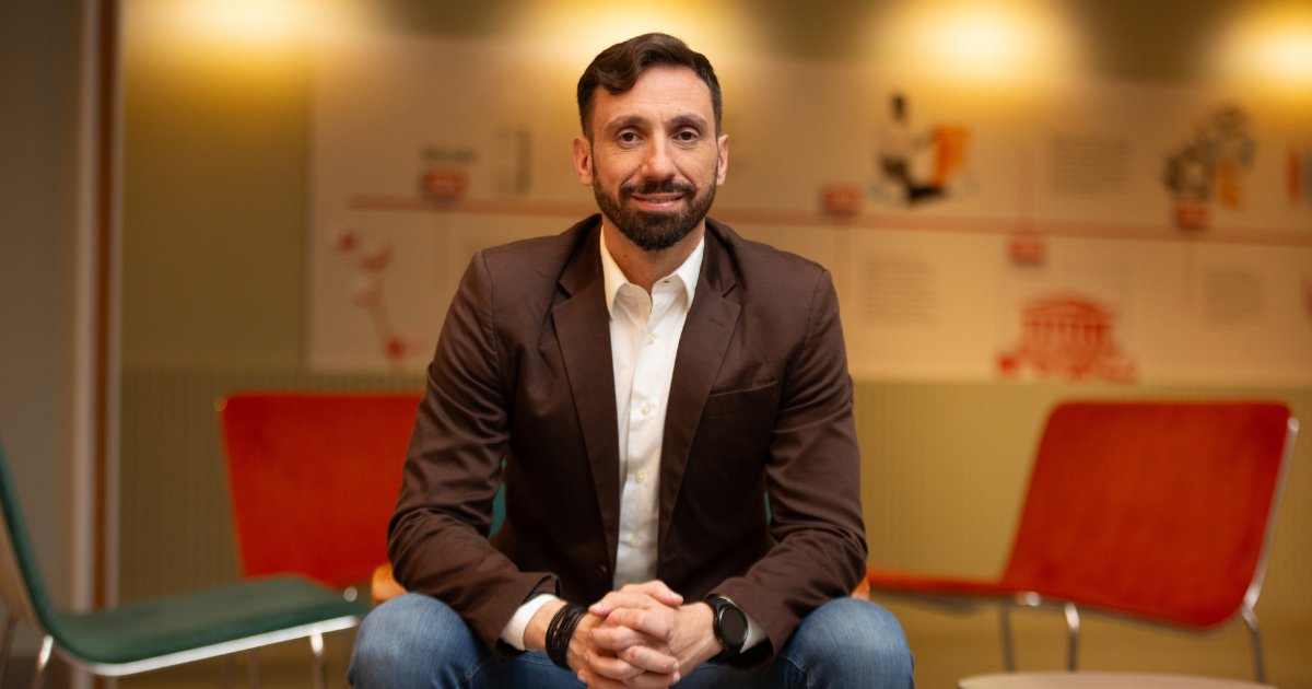 Imagem de Thiago Suzano, diretor de engenharia de software e dados da B3, sentado e sorrindo em um ambiente interno com iluminação suave. Ele veste um blazer marrom, camisa branca e calça jeans, e está com as mãos entrelaçadas à frente do corpo. Ao fundo, há cadeiras coloridas e um painel com ilustrações e textos desfocados.