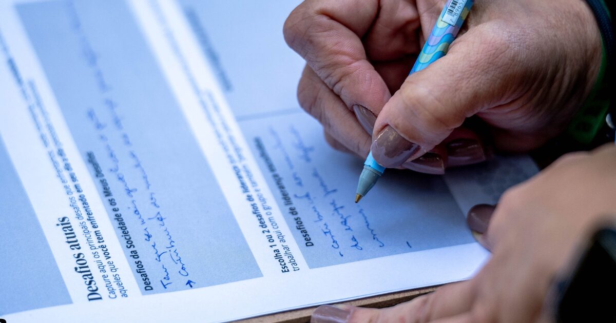 Imagem mostra uma mão com unhas pintadas de esmalte nude segurando uma caneta colorida enquanto escreve à mão em uma folha impressa. O documento possui títulos em negrito, como “Desafios atuais” e “Desafios da sociedade e os meus desafios”, sugerindo ser parte de uma atividade escrita reflexiva ou educativa. A imagem transmite um momento de participação ativa, possivelmente em um evento, oficina ou pesquisa (liderança de propósito)