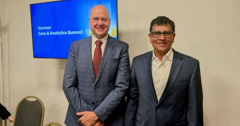 Imagem mostrando Jorg Heizenberg, vice-presidente e analista do Gartner, e Deepak Seth, diretor analista do Gartner, posando sorridentes em frente a uma tela que exibe o título "Gartner Data & Analytics Summit". Ambos estão vestidos formalmente com ternos; Jorg Heizenberg usa um terno azul com gravata vermelha, e Deepak Seth usa um terno azul escuro com camisa branca.