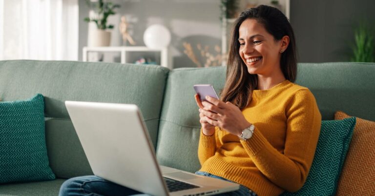 Mulher sorridente sentada em um sofá verde, usando um smartphone enquanto também está com um notebook no colo. Ela veste um suéter amarelo e parece estar em um ambiente doméstico moderno e aconchegante, sugerindo o uso da internet ou serviços digitais via smartphone em um momento de lazer ou trabalho remoto (brasil)