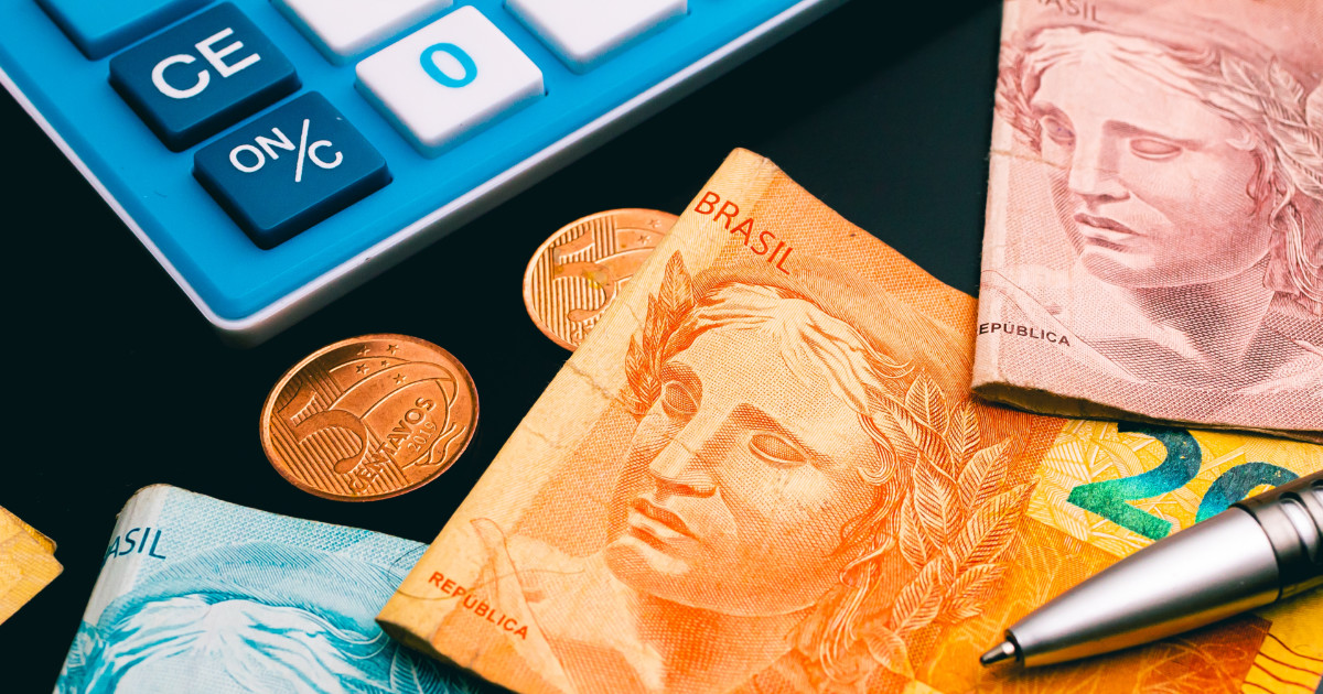 Imagem com cédulas e moedas de real, moeda oficial do Brasil, dispostas sobre uma superfície preta. Ao lado das cédulas e moedas, há uma calculadora azul e branca e uma caneta metálica prateada (reforma tributária, governo)