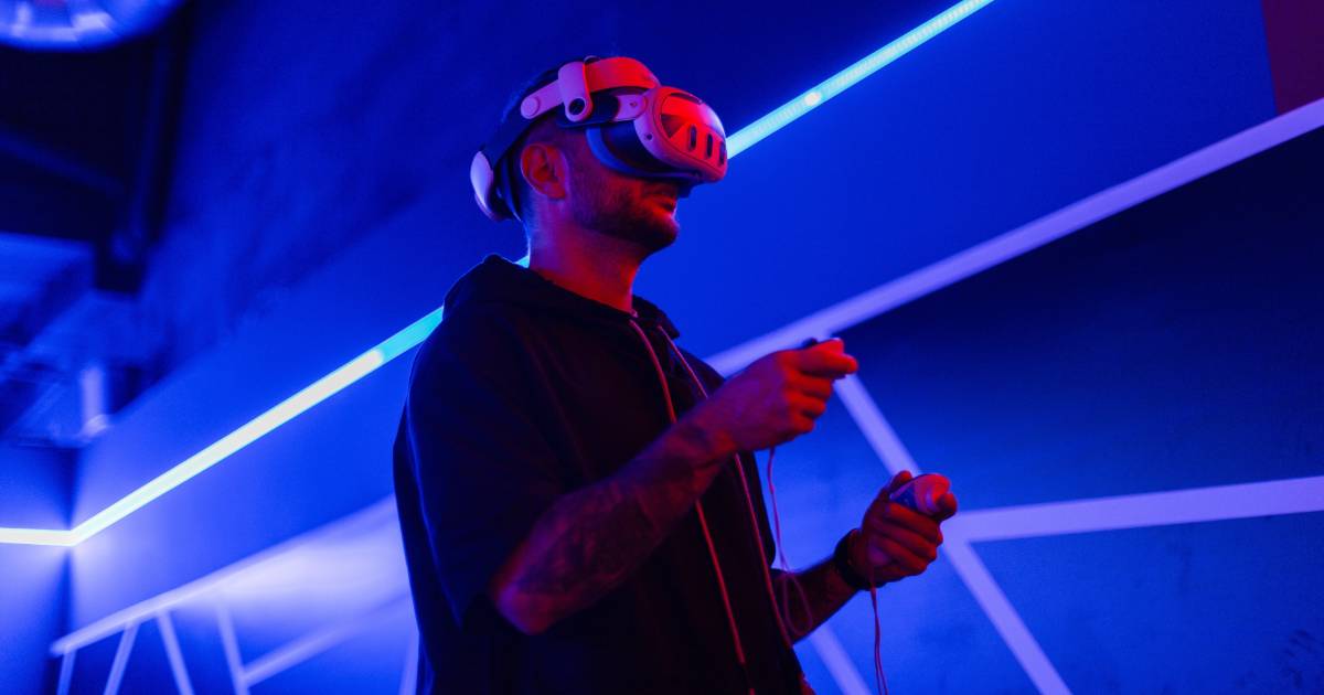 Homem utilizando um headset de realidade virtual com controles nas mãos em um ambiente escuro iluminado por luzes de LED azuis e roxas. Ele está concentrado na experiência imersiva, vestindo um moletom preto com capuz. O cenário tem um visual futurista com linhas geométricas nas paredes (Deloitte, computação espacial, hype)