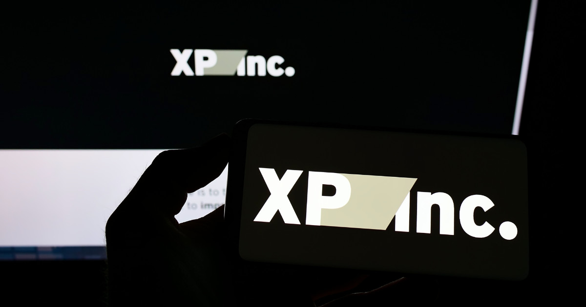 Logotipo da XP Inc. exibido em duas telas: uma em um smartphone segurado por uma mão em primeiro plano e outra ao fundo em um monitor, ambas com fundo escuro e letras brancas.