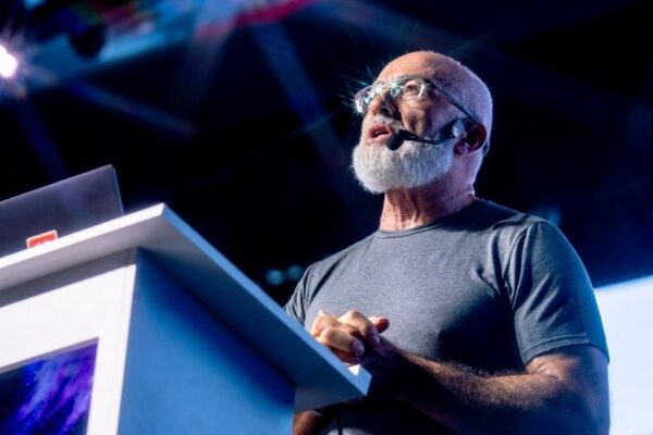 Um homem idoso com barba branca e óculos, vestindo uma t-shirt cinzenta, fala ao público com um microfone de cabeça enquanto está atrás de um púlpito branco com um portátil. A luz reflete nos seus óculos, criando um brilho marcante.