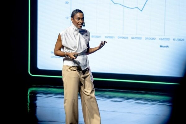 Uma mulher fala no palco com um microfone de cabeça e um controlo remoto na mão. Usa um top branco assimétrico e calças largas bege. Atrás dela, há um gráfico com dados e datas visíveis no ecrã digital.