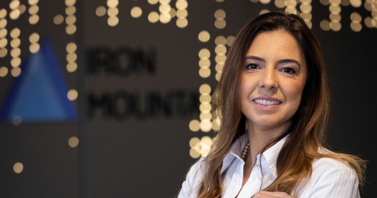 Ana Carla Martins Netto, diretora-geral comercial da Iron Mountain Brasil, sorri em frente ao logotipo da empresa. Ela veste camisa branca e colar de pérolas, com luzes desfocadas ao fundo criando um ambiente moderno e acolhedor.